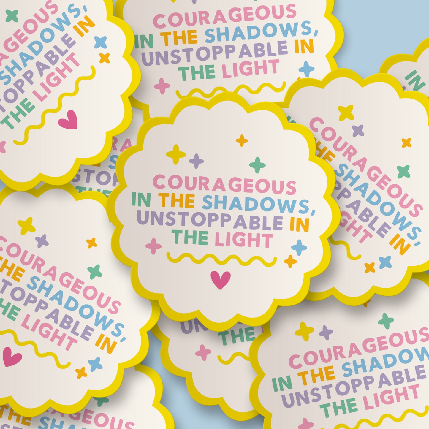 A pile of scalloped cloud-shaped stickers with a bright yellow border and the motivational quote 'Courageous in the shadows, unstoppable in the light' in pastel pink, blue, green, and purple lettering on a white background, set against a light blue backdrop.