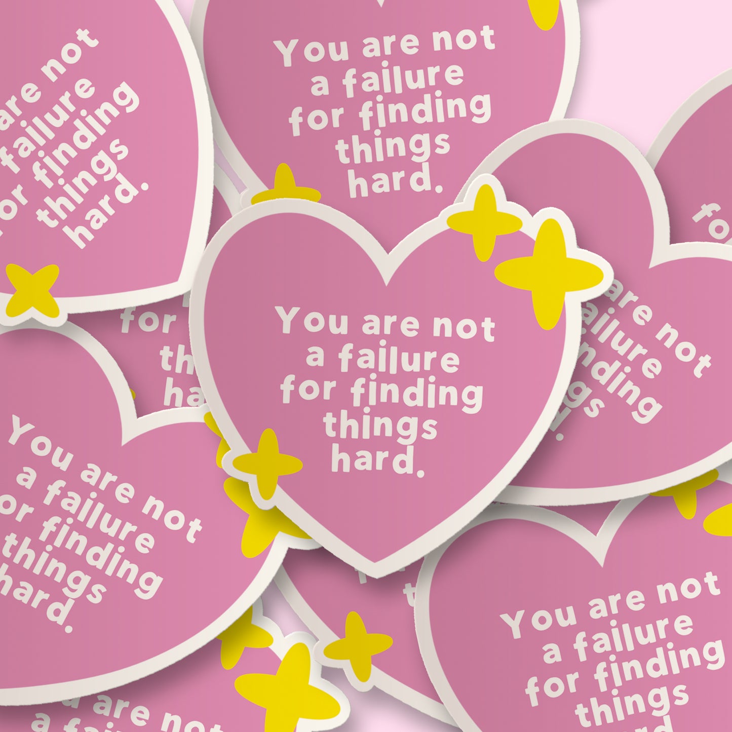 A close-up arrangement of pink heart-shaped stickers with white text reading 'You are not a failure for finding things hard.' and small yellow star accents, overlapping on a pale pink background.