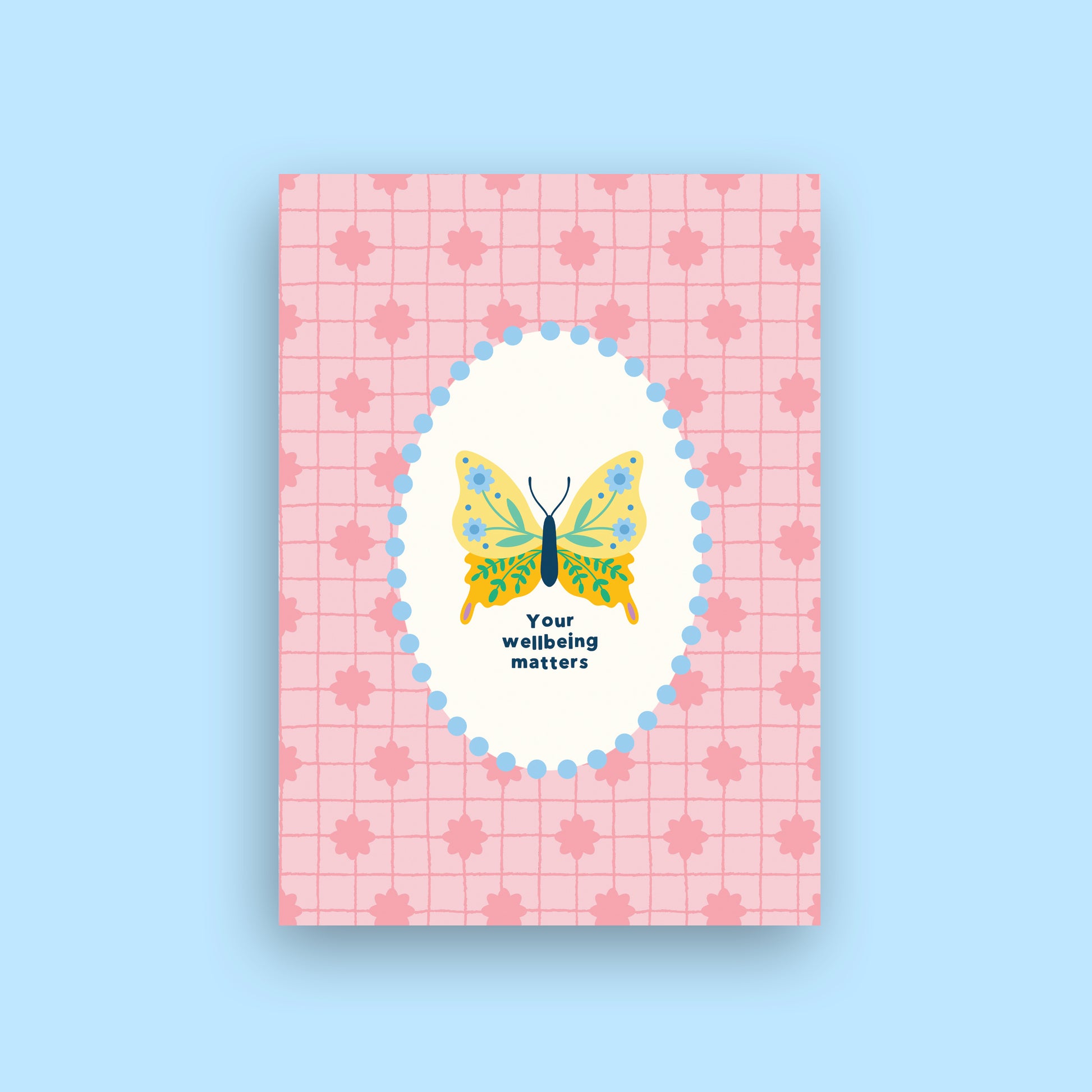 A rectangular pink card or notebook cover with a subtle grid and maple leaf pattern, featuring a central white oval with a colorful butterfly illustration and the text 'Your wellbeing matters' on a light blue background.