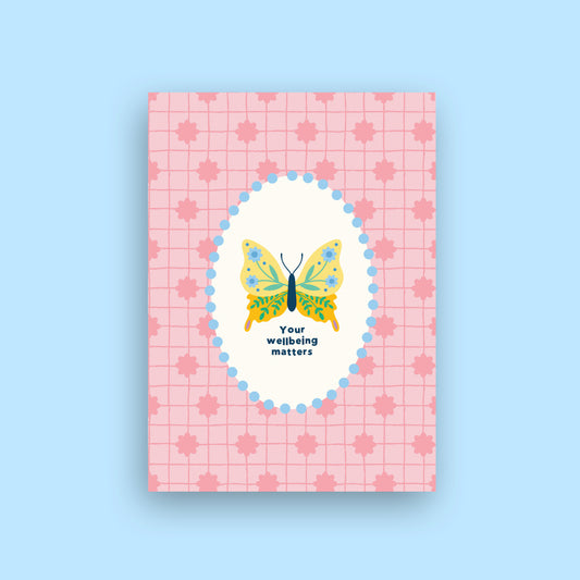 A rectangular pink card or notebook cover with a subtle grid and maple leaf pattern, featuring a central white oval with a colorful butterfly illustration and the text 'Your wellbeing matters' on a light blue background.